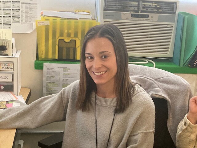 "Smiling professional sitting at a desk in an organized office, wearing a light sweater. An air conditioning unit and colorful files can be seen in the background, highlighting a productive work environment. Perfect for educational and office-related content."